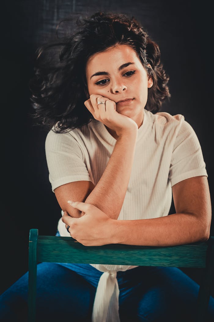 Portrait of a woman sitting pensively, wearing a casual white top. Moody, introspective atmosphere.